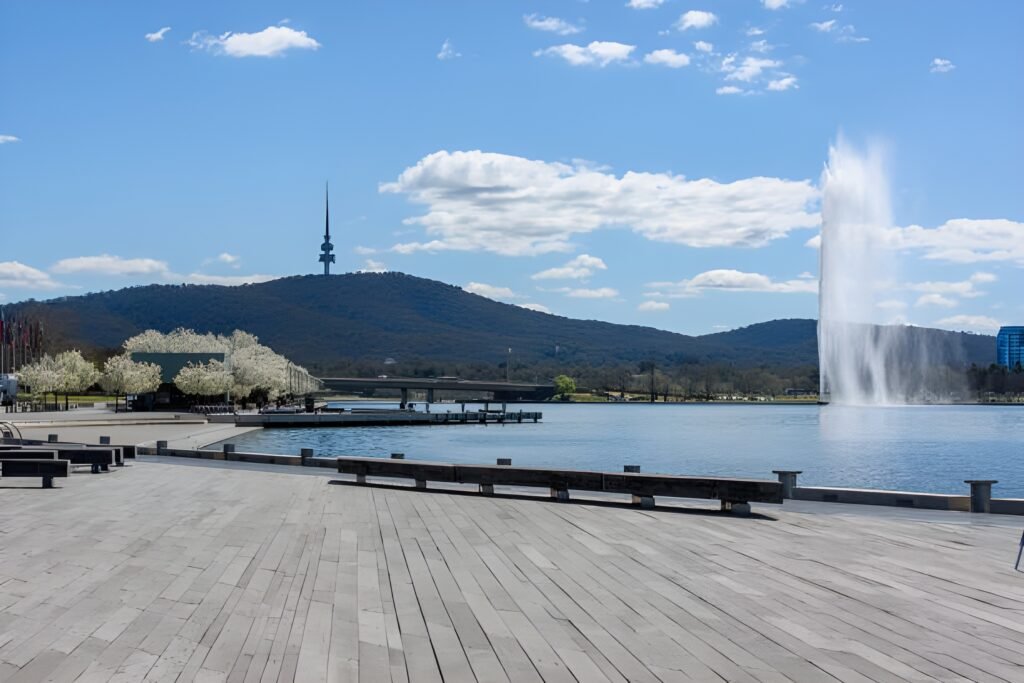 View of Lake Burley Griffin in Canberra with a fountain, surrounded by trees and hills in the background, under a clear blue sky.