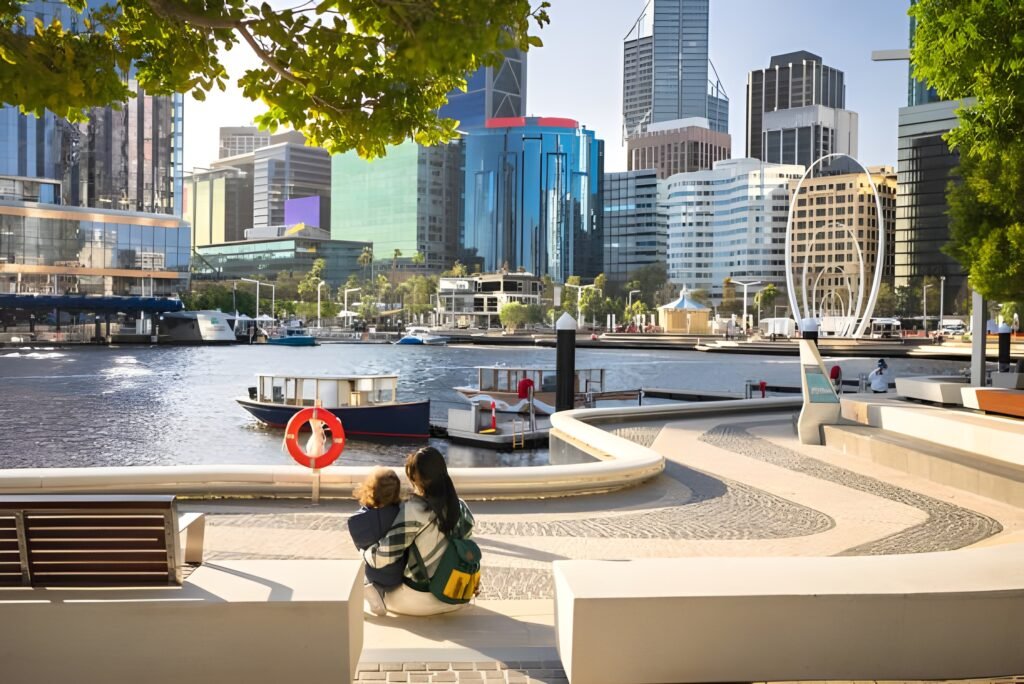 A couple sits on a bench by the waterfront in Perth, Australia, enjoying the view of the skyline and boats on the water.